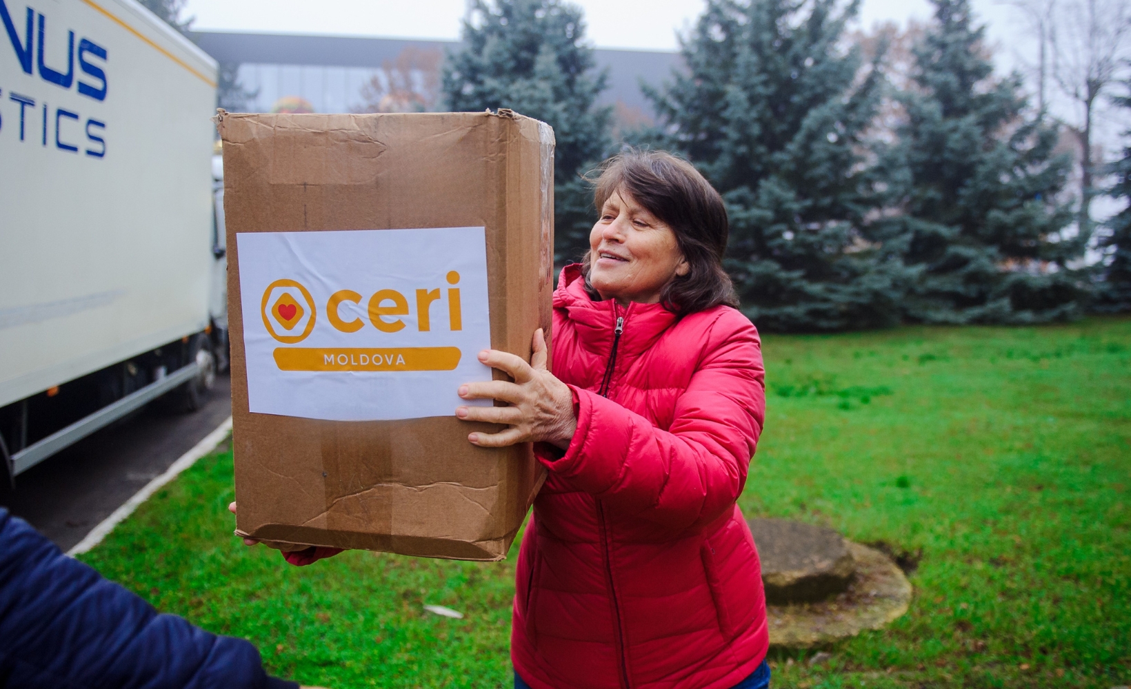 A woman in a red jacket holds a large cardboard box labeled "CERI Moldova" outdoors near evergreen trees and a logistics truck.