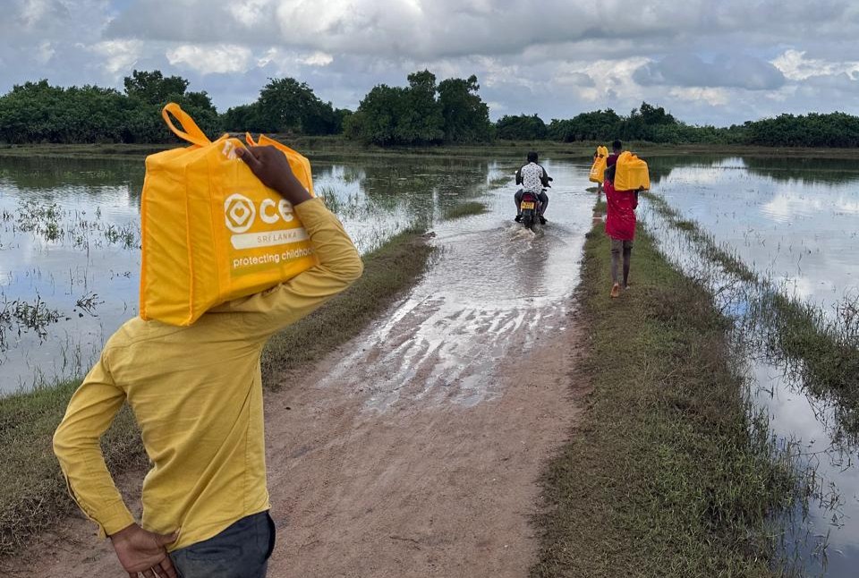 Three people carrying yellow bags labeled "CERI Sri Lanka" walk on a muddy path through a flooded area.