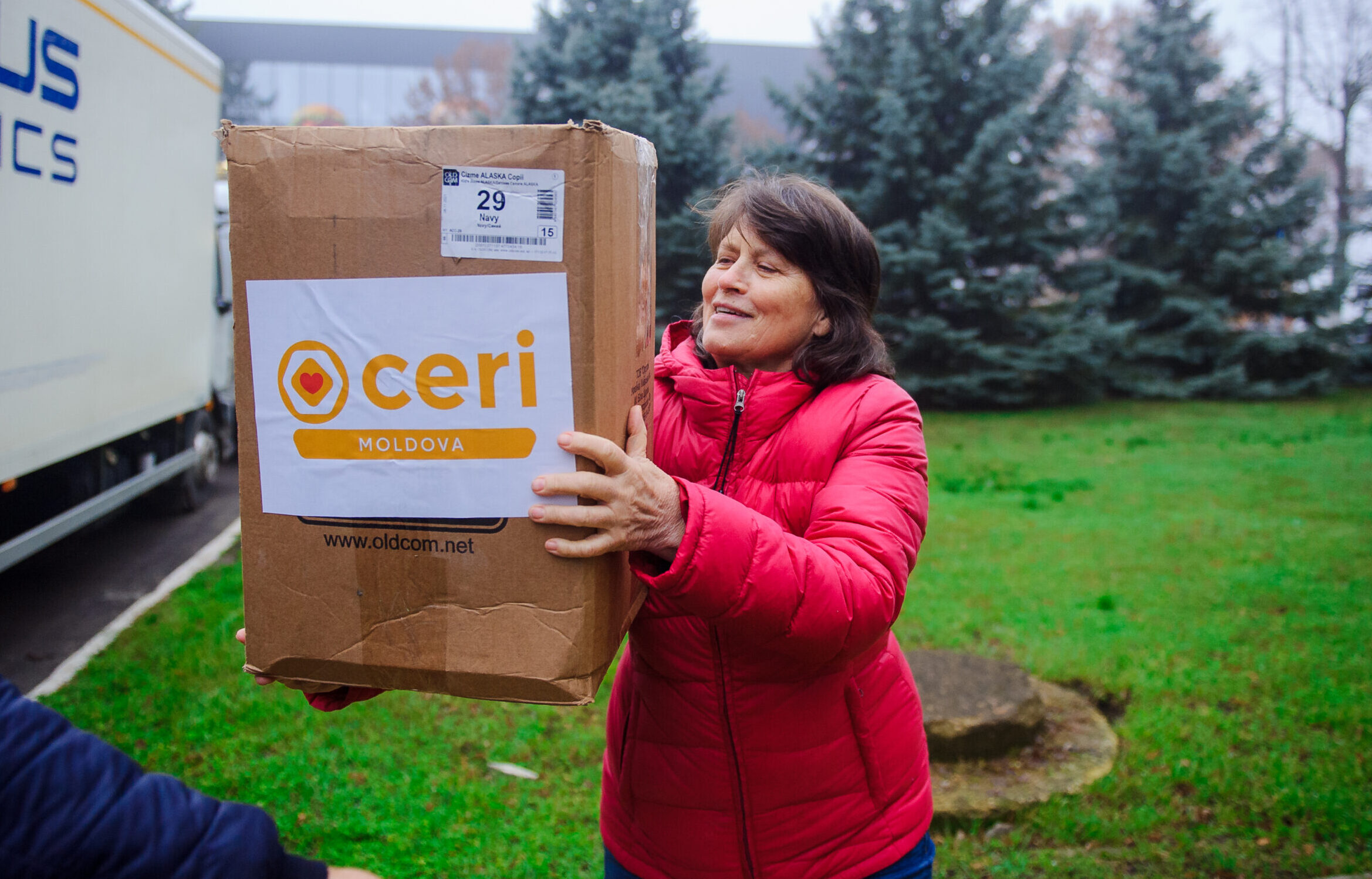A woman in a red jacket holds a large cardboard box labeled "CERI Moldova" outdoors near evergreen trees and a logistics truck.