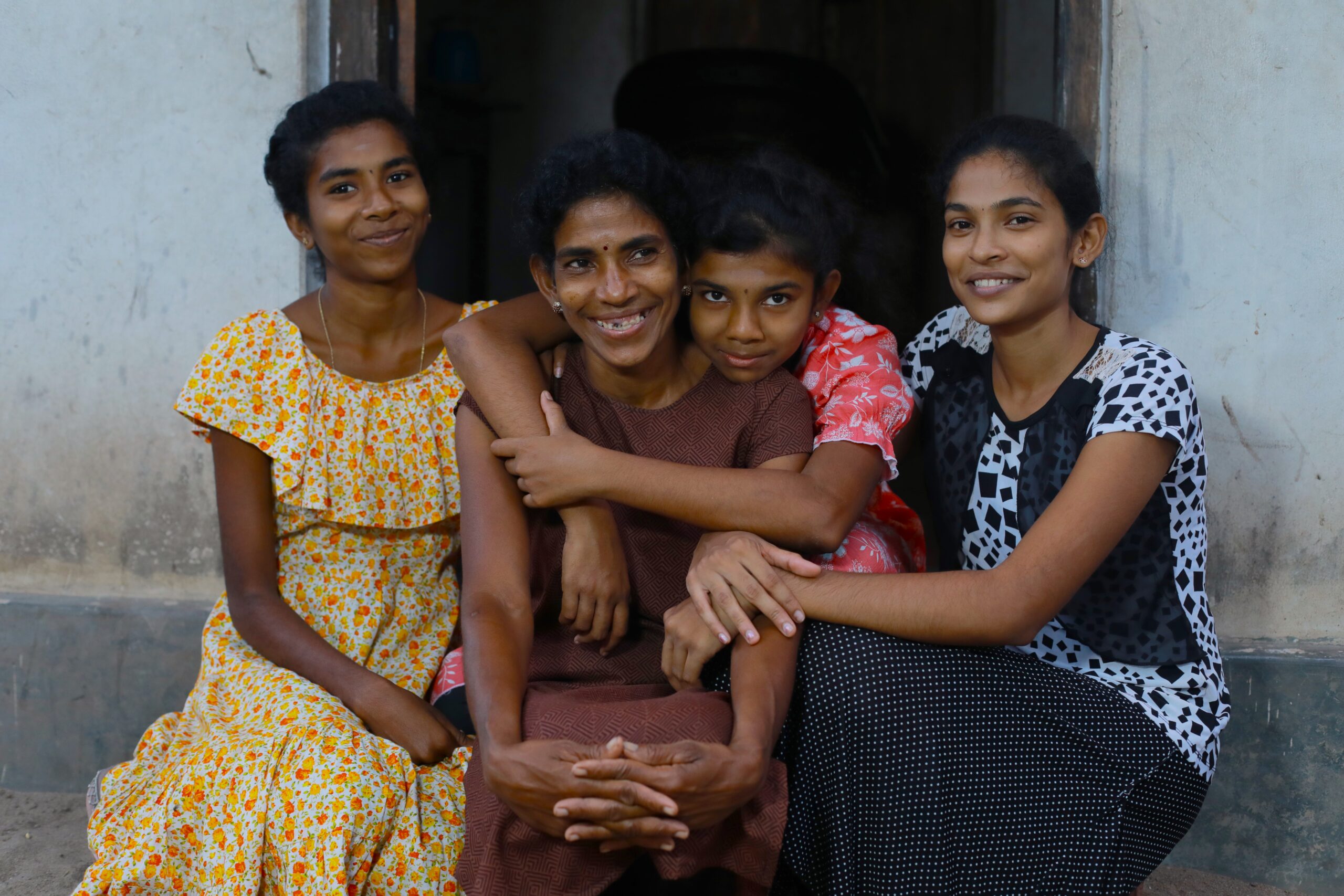 Four people sit closely together in front of a doorway, smiling and facing the camera. The group includes three young women and one older woman in the center.